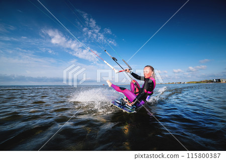 A young Caucasian woman in a wetsuit rides a kiteboard. An athlete performs a trick while sitting on a board with a kissing grimace. Beauty and grace in water sports A young Caucasian woman in a wetsuit rides a kiteboard. An athlete performs a trick while sitting on a board with a kissing grimace. Beauty and grace in water sports 115800387