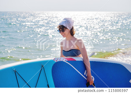 Young athletic woman stands on the beach and surfs on a SUP. The girl holds a board and goes to the sea to swim 115800397