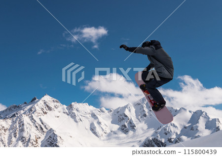Portrait of a young man snowboarder jumping on a snowboard in sportswear, against the backdrop of high snow-capped mountains and sky 115800439