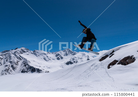 sporty male tourist snowboarding in the sunny Alps performs a trick of grabbing the board and flying off a kicker or big air. Professional snowboarder performing tricks sporty male tourist snowboarding in the sunny Alps performs a trick of grabbing the board and flying off a kicker or big air. Professional snowboarder performing tricks 115800441
