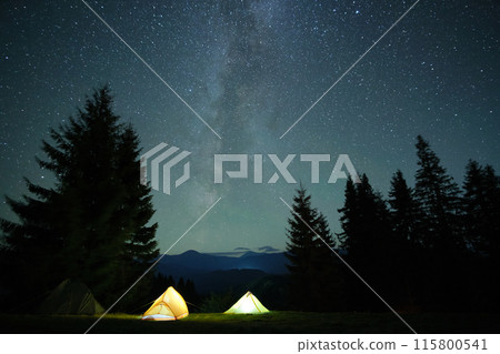 Bright illuminated tourist tents glowing on camping site in dark mountains under night sky with sparkling stars. Active lifestyle concept. 115800541