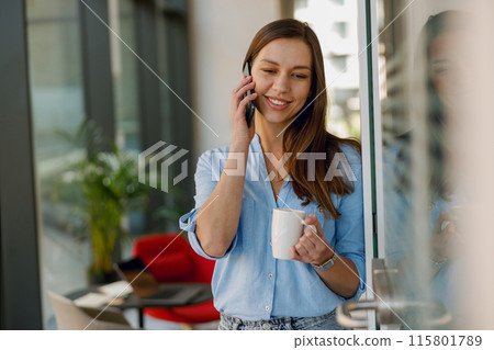 A young woman in a modern office chats on her phone, looking relaxed and professional with a coffee A young woman in a modern office chats on her phone, looking relaxed and professional with a coffee 115801789