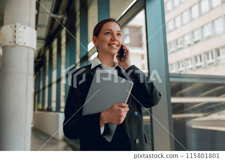 Confident businesswoman in a modern office talking on the phone and holding documents Confident businesswoman in a modern office talking on the phone and holding documents 115801801