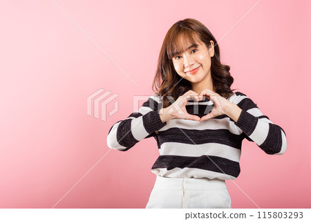 A confident woman smiles and creates a heart symbol with two hands on a pink background. Asian portrait of a beautiful young female expressing love and happiness for Valentine's Day. 115803293