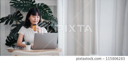 A young woman in a white shirt working on a laptop at home, holding a coffee cup, with a green plant in the background. Ideal for themes of remote work, freelance, and modern home office. A young woman in a white shirt working on a laptop at home, holding a coffee cup, with a green plant in the background. Ideal for themes of remote work, freelance, and modern home office. 115806283