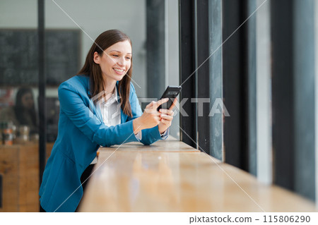 A professional woman in a blue blazer smiles while using her smartphone in a modern office with large windows and natural light. 115806290