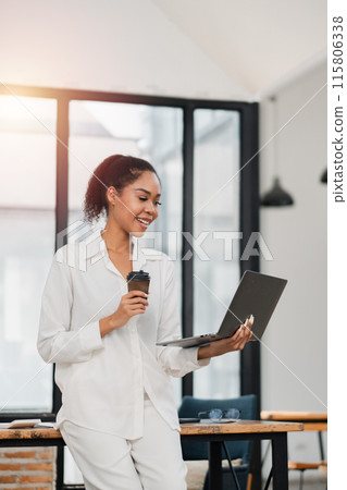 Young woman in white attire holding a coffee cup and laptop in a bright, modern office with large windows. 115806338
