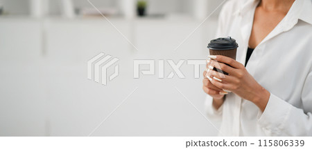 Close-up of a woman holding a coffee cup in a modern minimalist setting, wearing a white shirt with manicured nails. 115806339