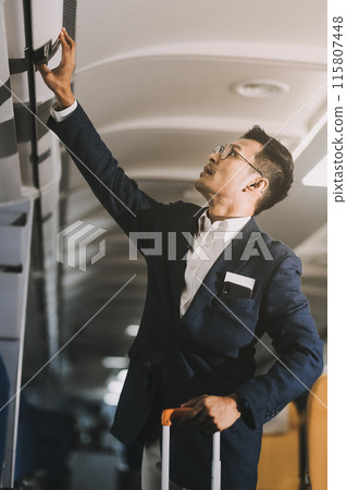 Handsome Asian male passenger takes his carry-on luggage out of the overhead locker after landing at his destination. transportation concept Handsome Asian male passenger takes his carry-on luggage out of the overhead locker after landing at his destination. transportation concept 115807448