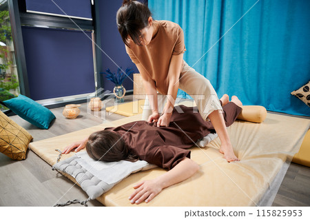 Masseuse making thai yoga massage. Woman therapist doing traditional massage treatment for female patient using fists. Client lying on mat, meditating, enjoying hand massage in calm atmosphere. Masseuse making thai yoga massage. Woman therapist doing traditional massage treatment for female patient using fists. Client lying on mat, meditating, enjoying hand massage in calm atmosphere. 115825953