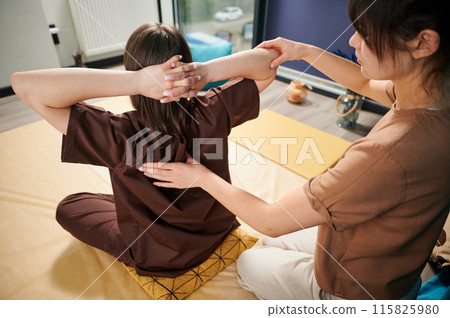 Masseuse making thai yoga massage. Woman therapist doing traditional massage treatment for female patient indoors. Back view of client sitting on mat, meditating, enjoying hand massage. 115825980