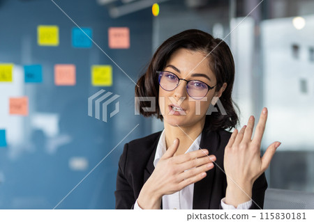 Confident businesswoman presenting and explaining ideas during a video call in a modern office with sticky notes in the background. 115830181