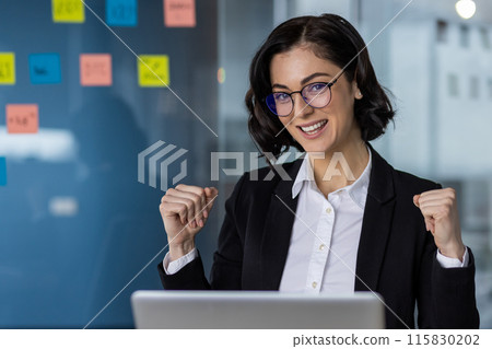 Happy businesswoman in formal attire celebrating a success with fists raised, working on laptop in modern office. Happy businesswoman in formal attire celebrating a success with fists raised, working on laptop in modern office. 115830202