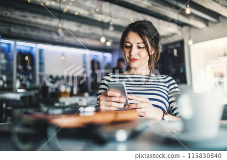 Woman working remotely from coffee shop. Cappucino and laptop on table. Female freelancer scrolling on smartphone in cafe. 115830840