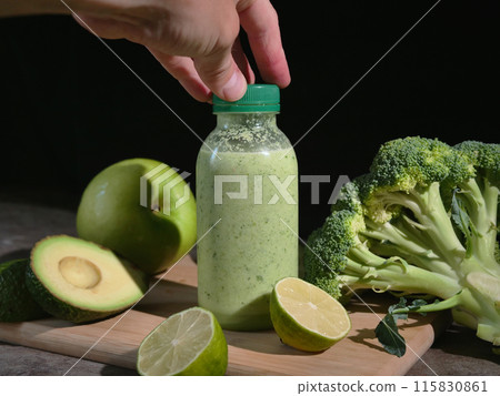 Refreshing green smoothie in bottles on wooden board Hand picks up smoothie from table with avocado, lime, broccoli, apple, cucumber. Perfect for healthy lifestyle content. Ideal smoothie  115830861