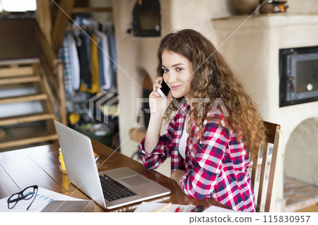 A beautiful university student is writing her thesis on a laptop, sitting at home at table and making phone call. A beautiful university student is writing her thesis on a laptop, sitting at home at table and making phone call. 115830957