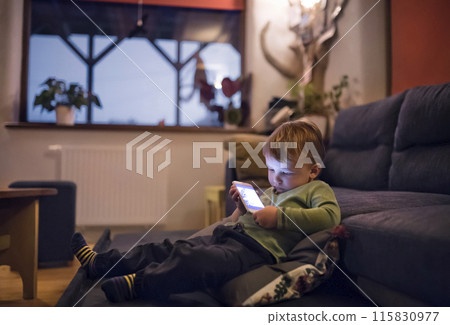 Cute boy sitting on bed watching something on smartphone. Children's screen time. 115830977