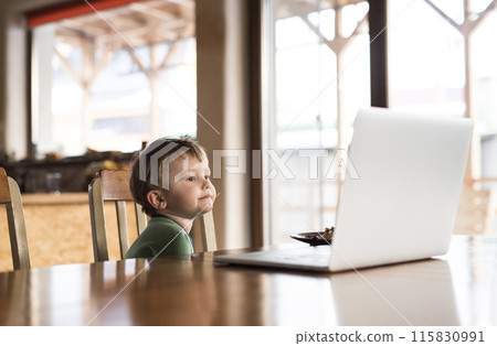 Little boy watching cartoons on laptop, sitting at table and eating porridge. 115830991