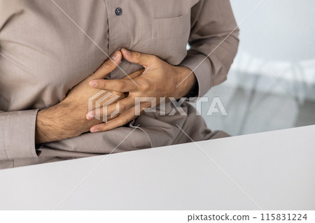 Close-up image of a man holding his stomach with both hands, indicating pain or discomfort. The image conveys concepts of stomach ache, illness, anxiety, and health issues. 115831224