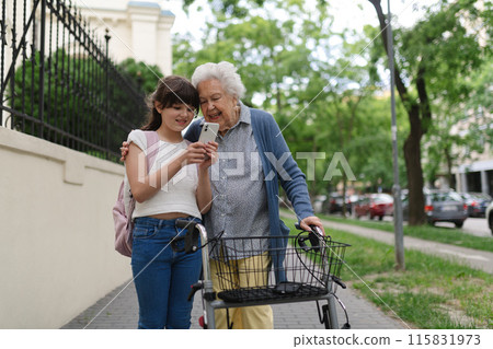Grandma picking up young girl from school at afternoon. Granddaughter showing something on smartphone to senior grandmother, teaching her how to work with technology. Grandma picking up young girl from school at afternoon. Granddaughter showing something on smartphone to senior grandmother, teaching her how to work with technology. 115831973