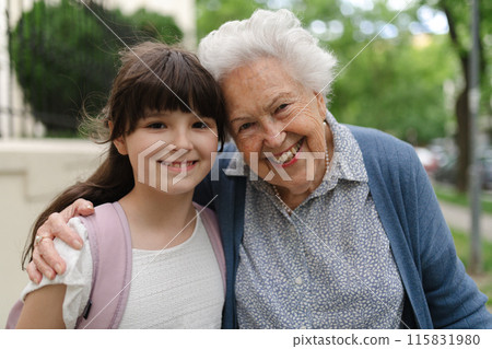 Grandma picking up young girl from school at afternoon. Granddaughter spending time with senior grandmother outdoors in city after school. 115831980