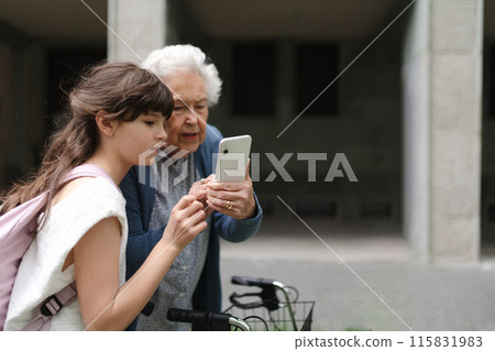 Grandma picking up young girl from school at afternoon. Granddaughter showing something on smartphone to senior grandmother, teaching her how to work with technology. Grandma picking up young girl from school at afternoon. Granddaughter showing something on smartphone to senior grandmother, teaching her how to work with technology. 115831983