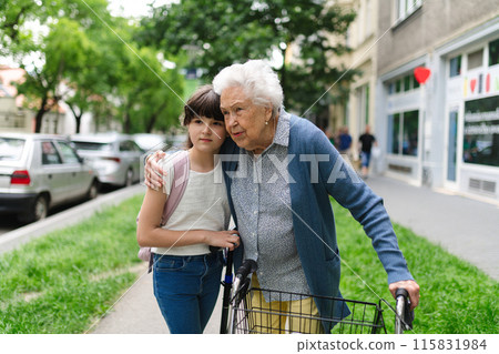 Grandma with walker picking up young girl from school at afternoon. Granddaughter spending time with senior grandmother outdoors in city after school. 115831984