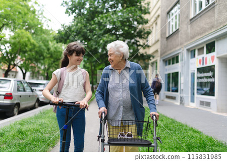 Grandma with walker picking up young girl from school at afternoon. Granddaughter spending time with senior grandmother outdoors in city after school. Grandma with walker picking up young girl from school at afternoon. Granddaughter spending time with senior grandmother outdoors in city after school. 115831985