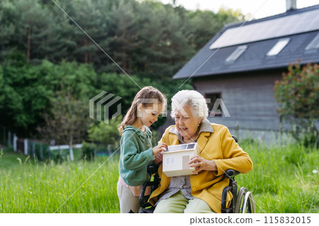 Granddaughter showing grandma model of house with solar panels on roof. Senior lady in wheelchair spending time in nature. 115832015