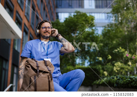 Doctor sitting on low concrete wall, listening music, wearing headphones. Winding down from busy workday in hospital. 115832083
