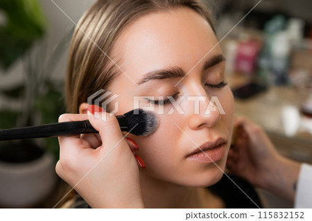 Young lady comes to beauty salon to do professional makeup. Makeup artist lightly applies highlighter with special brush on client face 115832152