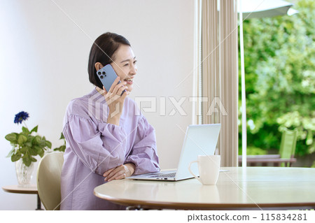 A middle-aged woman looking at a computer while using her smartphone in the living room 115834281