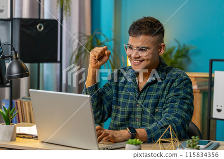 Overjoyed Indian man working on laptop celebrate success win money in lottery get online good news 115834356