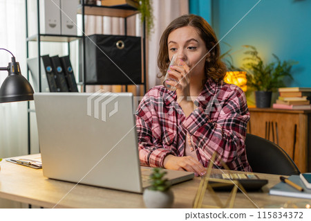 Businesswoman girl sitting at workplace desk drinking water while working with laptop in home office Businesswoman girl sitting at workplace desk drinking water while working with laptop in home office 115834372