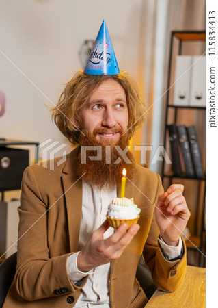 Happy business office man wears festive birthday hat hold cupcake makes wish blowing burning candle Happy business office man wears festive birthday hat hold cupcake makes wish blowing burning candle 115834413