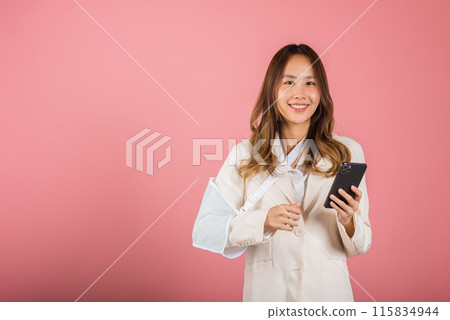 Woman confident smiling broken arm after accident and wear arm splint for treatment and hold smartphone, happy Asian female sling supported hand isolated on pink background, social media, copy space 115834944