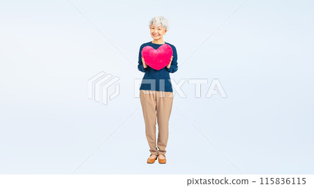 Full body photo of a senior woman holding a heart-shaped cushion 115836115