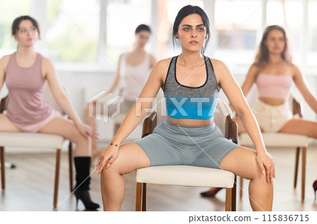 Slender athletic young women perform various dance movements with legs while sitting on chairs 115836715