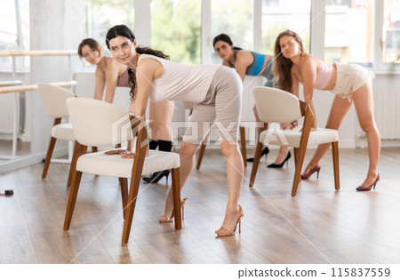 Leaning on chair seat with hand foot, young woman performs lap dance during group workout. 115837559