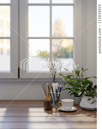 A wooden table by the window in a room features a coffee cup, a reed diffuser, and decorative plants 115838924