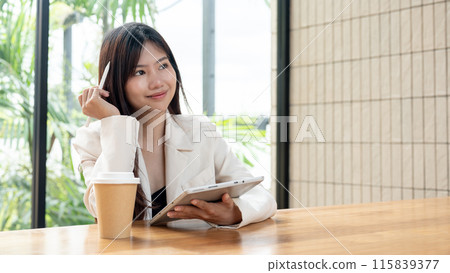 An attractive, thoughtful Asian businesswoman is working remotely from a cafe, using her tablet. An attractive, thoughtful Asian businesswoman is working remotely from a cafe, using her tablet. 115839377