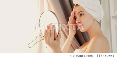 Morning care. Product applying. Facial skin preparation. Daily beauty routine procedure. Smiling young woman touching face checking looks in mirror free space. 115839664