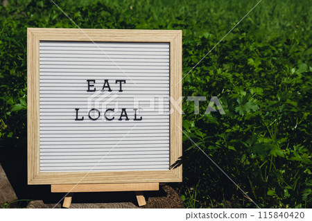 Letter board with text EAT LOCAL on background of garden bed with green herb parsley. Organic farming, produce local vegetables concept. Supporting local farmers 115840420