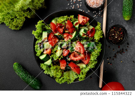 Vegetable salad of tomatoes, cucumbers, onions, lettuce dressed with aromatic dressing in a black plate on a gray background 115844322