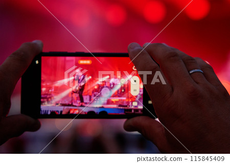 Person standing in front of stage, recording live music concert on smartphone 115845409