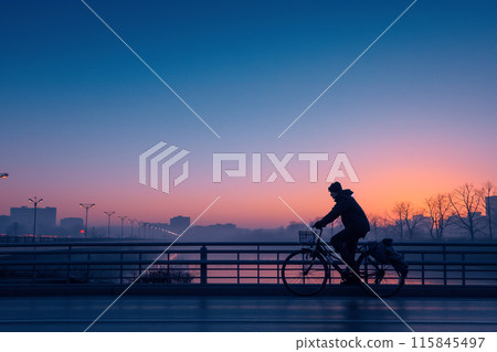 Man biking across bridge at sunrise with city skyline in background 115845497