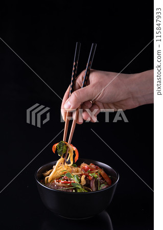 Hand using chopsticks pickup instant noodles with smokes isolated on black background. 115847933