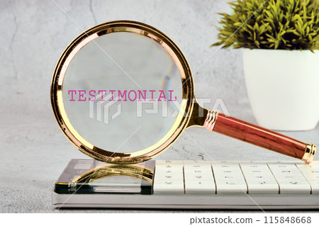 Business concept. TESTIMONIAL is written through a magnifying glass standing on a calculator on a gray background with a plant in the background without focus 115848668