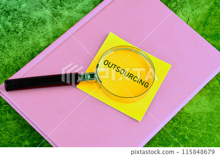 Copy space. Business concept. OUTSOURCING symbol written through a magnifying glass on a yellow sticker lying on a business notebook on a green background 115848679