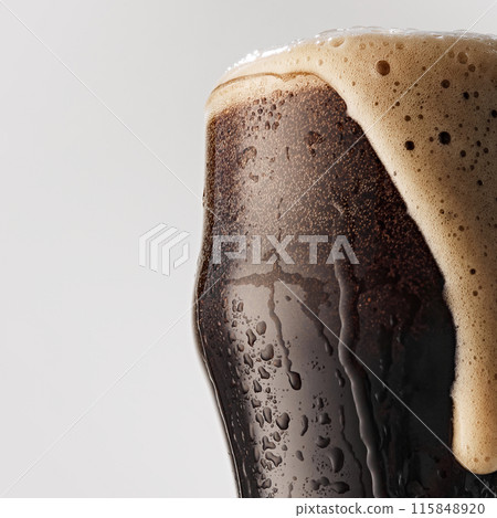 Close-up of pint glass of dark beer, with a creamy, overflowing head of foam against grey studio background. 115848920